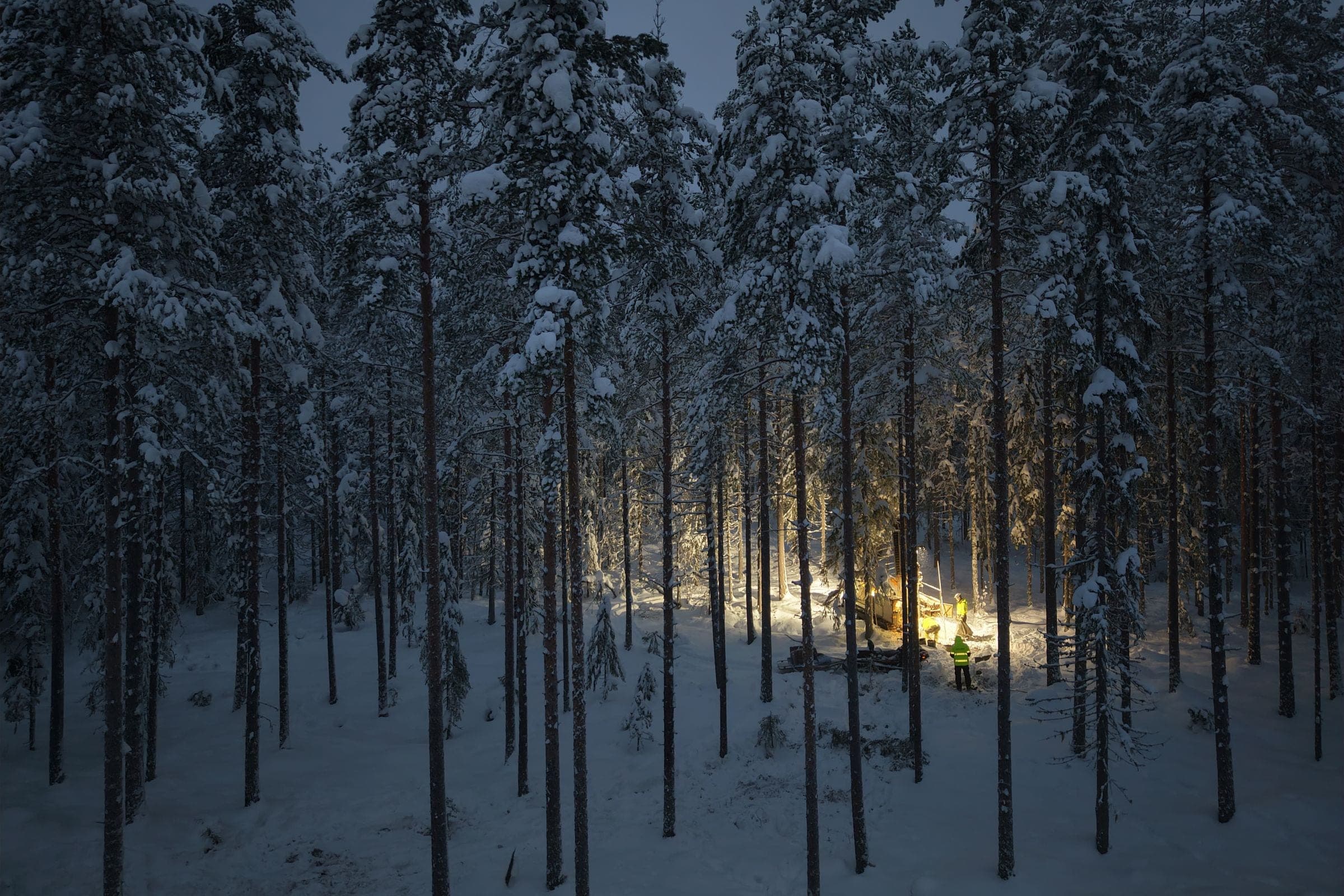 A lit core drilling rig and crew in a snow-covered pine forest at dusk.